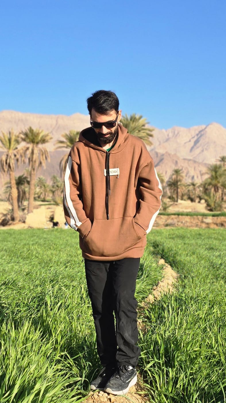 ایستاده در دشتی سبز زیر کوه‌های بیابانی و نخل‌ها
